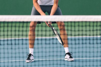 A tennis player's legs visible behind a net, with his posture clearly hunched forward as if about to hit an incoming ball.