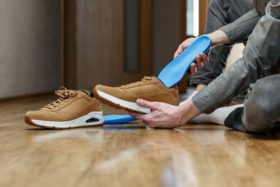 Man fitting blue orthotic insoles into brown trainers while sitting on the wooden floor