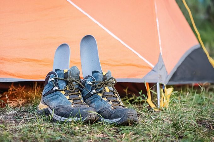  A pair of hiking boots with insoles standing upright inside of them, placed next to an orange tent on a patch of grass.